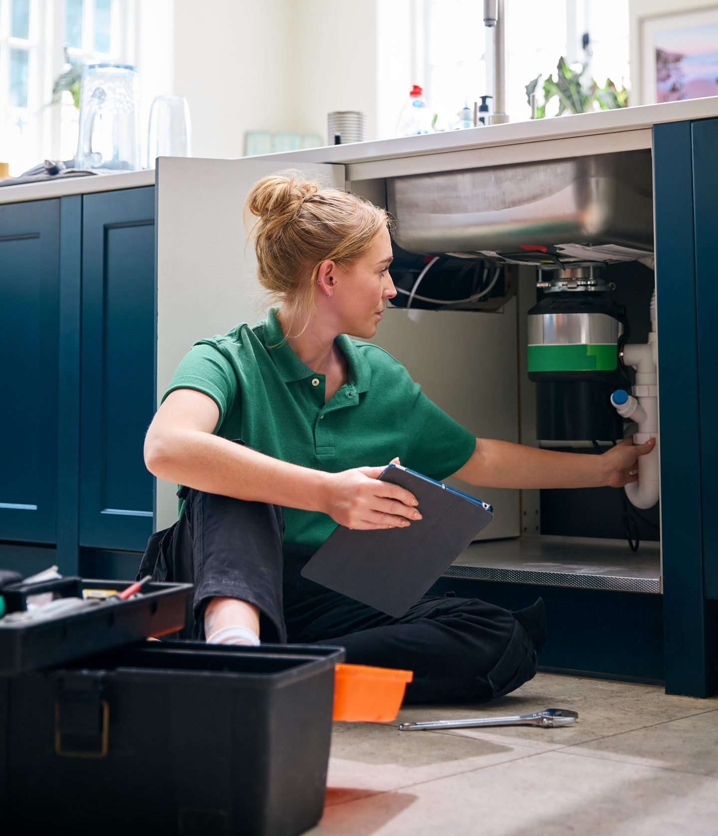 Female Plumber With Digital Tablet Fixing Waste Disposal Unit In Domestic Kitchen Sink