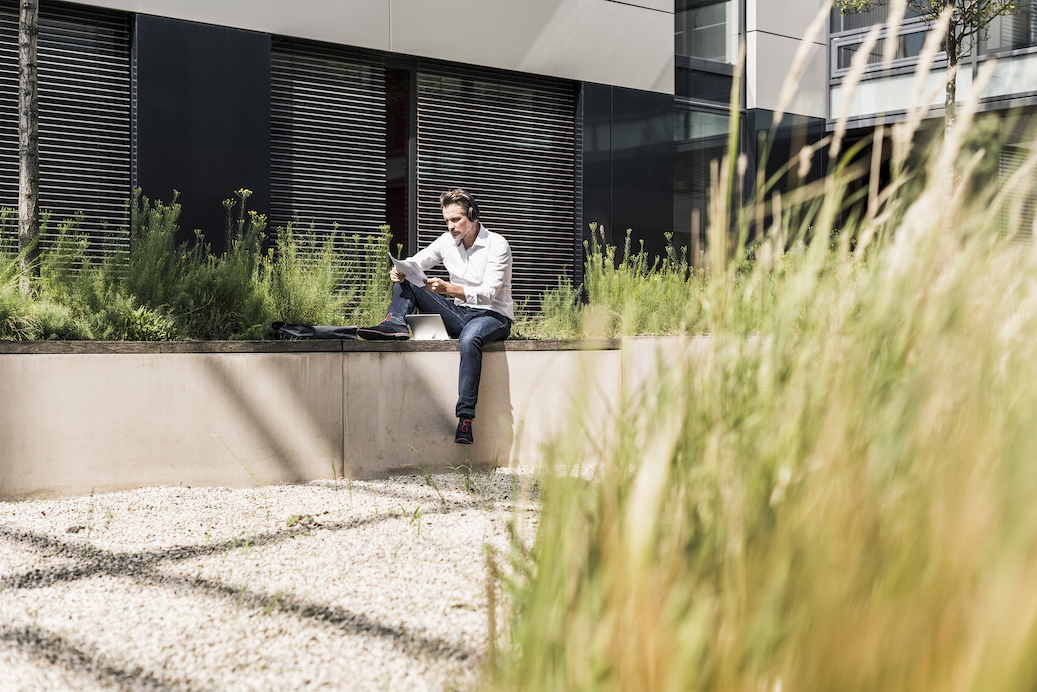 Businessman with headphones sitting on wall outside office building reading document