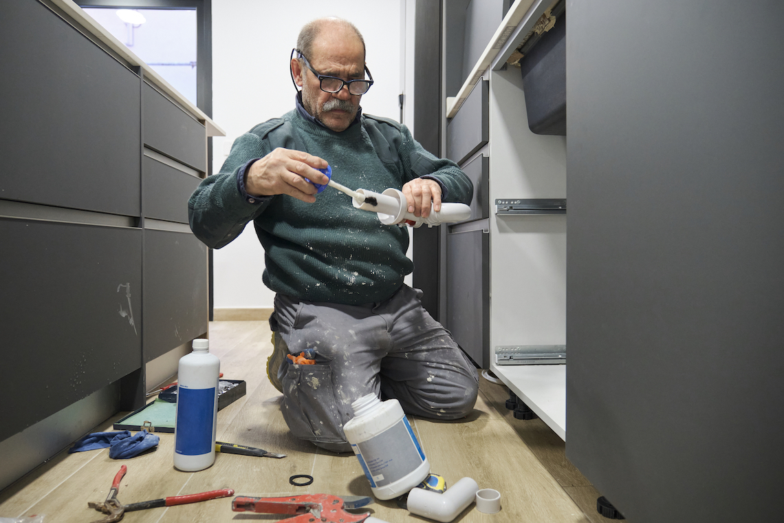 Mature plumber applying glue to a sink siphon white plastic pipe.